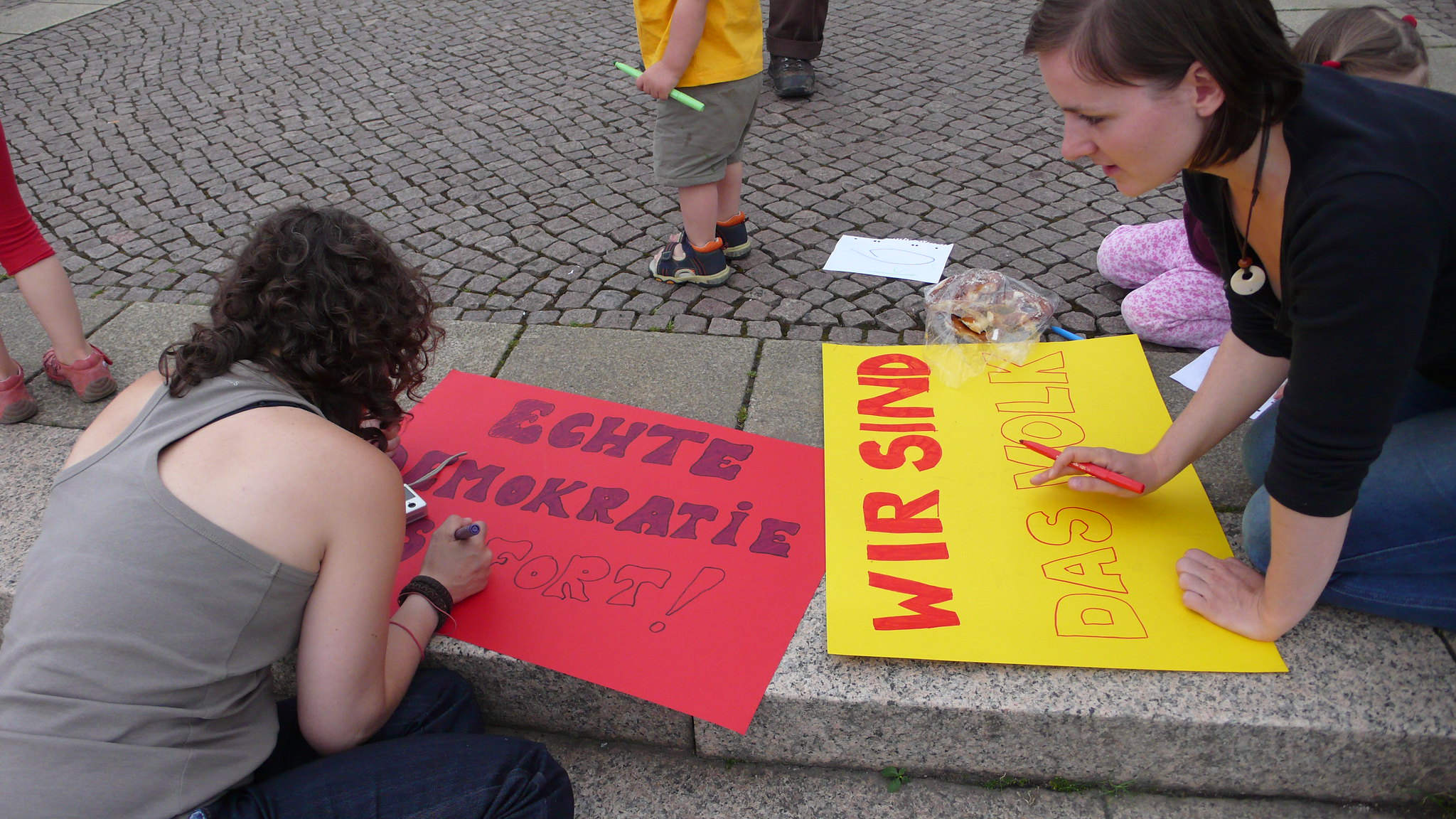 Zwei Frauen gestalten Plakate mit den Aufschriften „Echte Demokratie sofort!“ und „Wir sind das Volk“